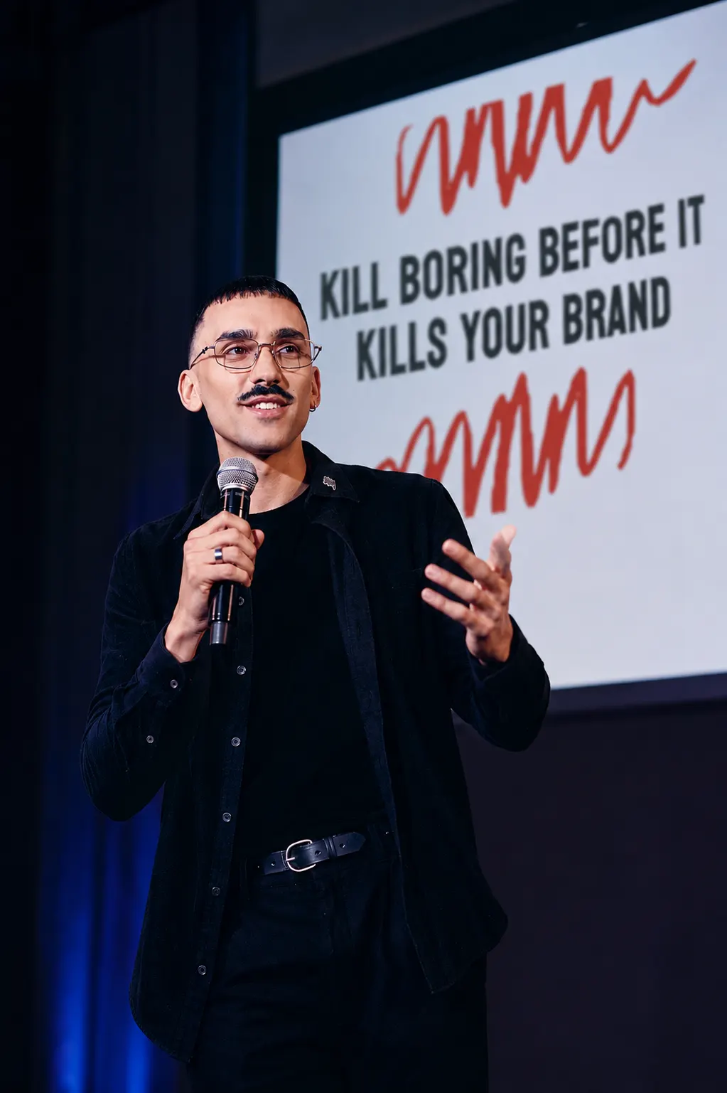 Marcus Willis speaking on stage, with "Kill boring before it kills your brand" on screen behind him